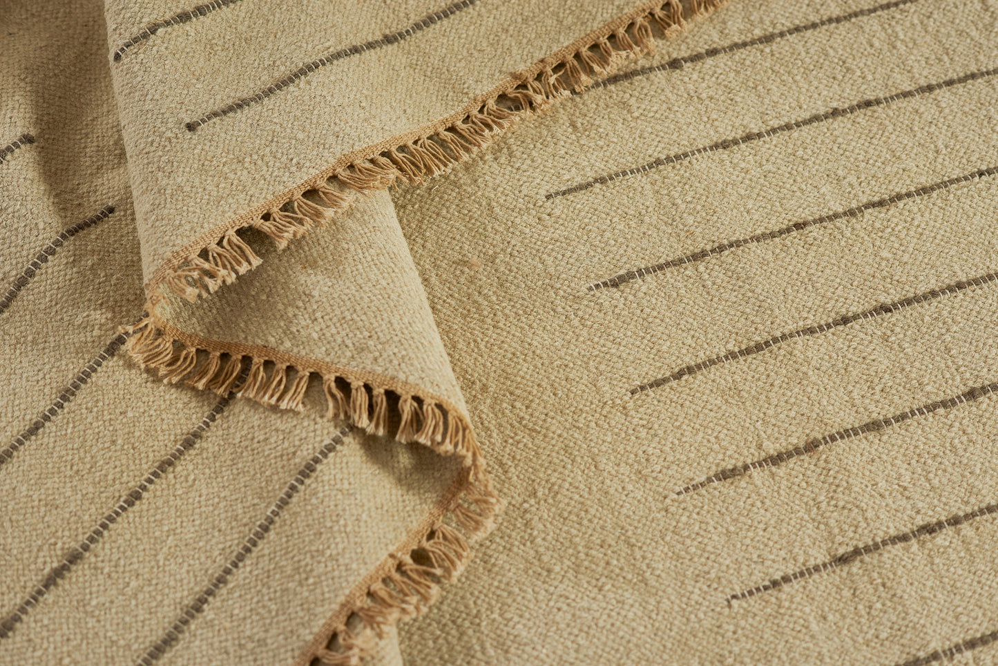 Beige woven rug with slim dark stripes and fringed edges, coastal casual floor decor.