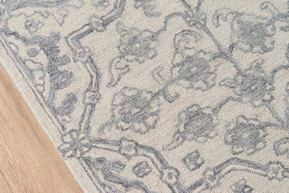 Grey damask-pattern area rug with ornate border on a light background, partially on a wooden floor.