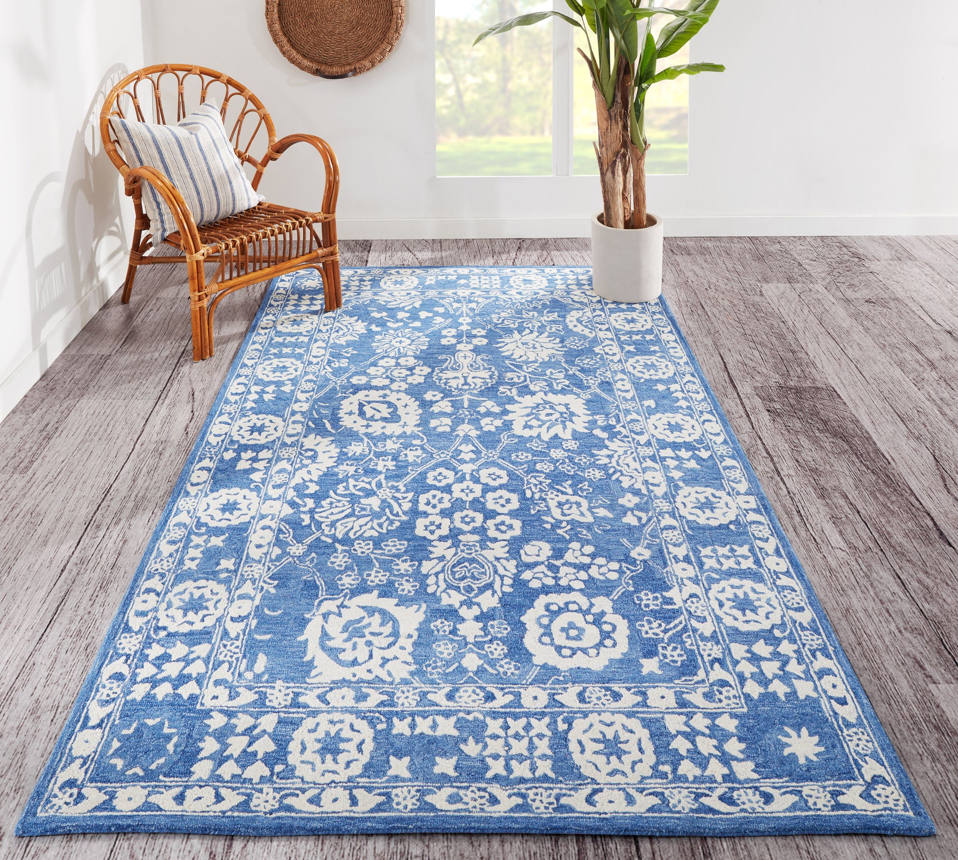 Rectangular blue rug with white floral medallion border pattern; woven look, in bright living space with wicker chair and plant.