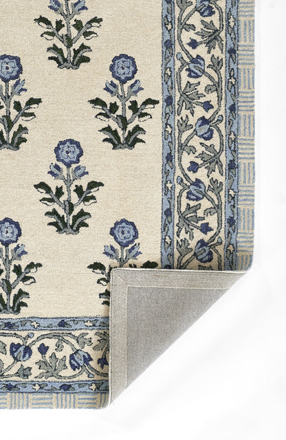 Beige woven rug with blue floral motifs and ornate border; folded corner shows backing.