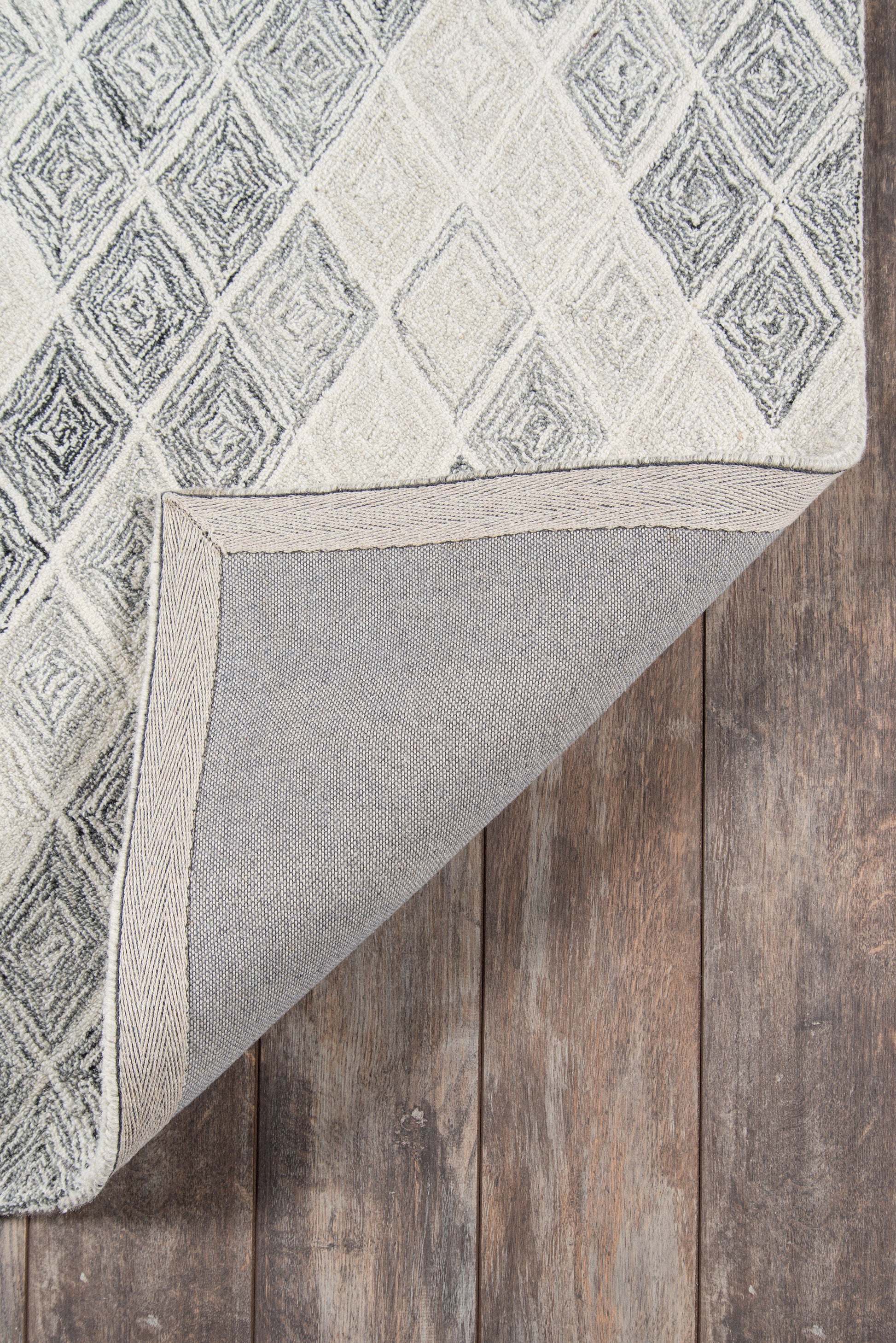 Grey geometric diamond rug with light and dark tones, folded corner reveals backing on wooden floor.