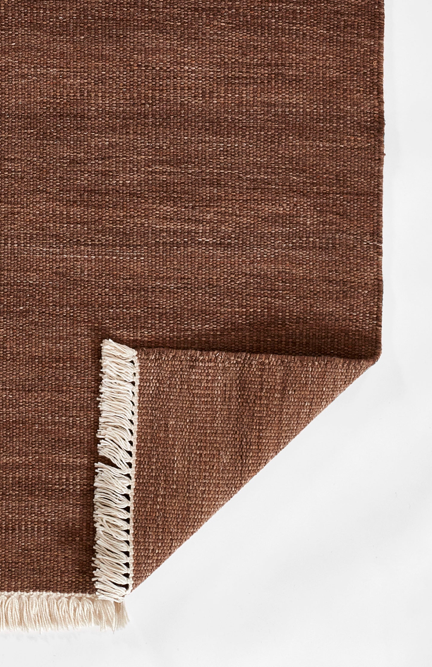 Brown woven rug with beige fringe along the edge; flat-weave texture with a folded corner.