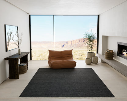 Minimalist living room with floor-to-ceiling window, tan bean bag, dark rug, rustic wood console, plants, and fireplace.
