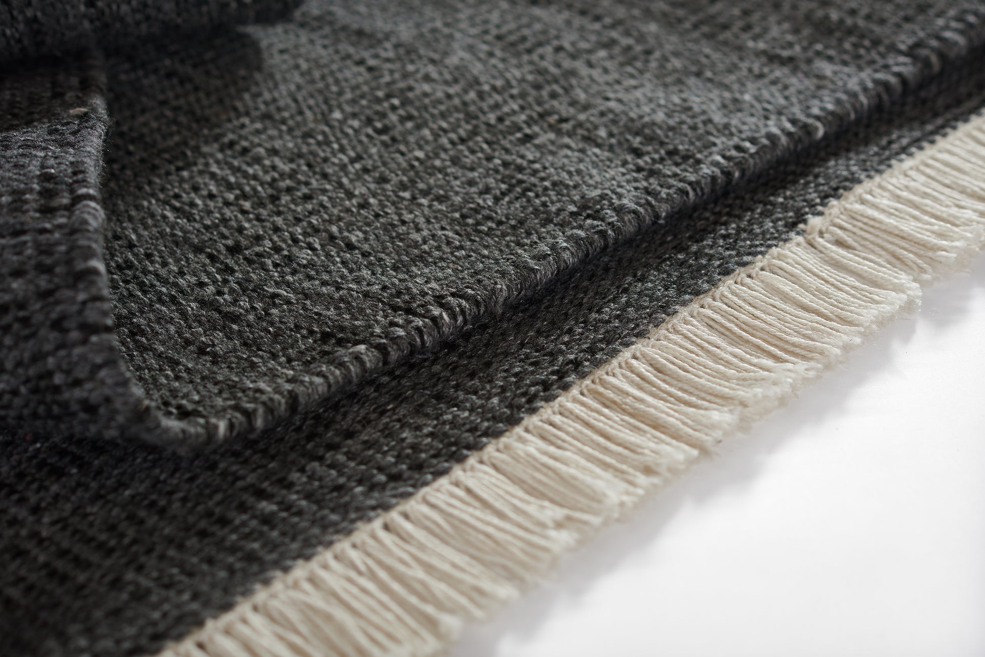 Charcoal woven rug with light beige fringe along edge; close-up shows textured weave.