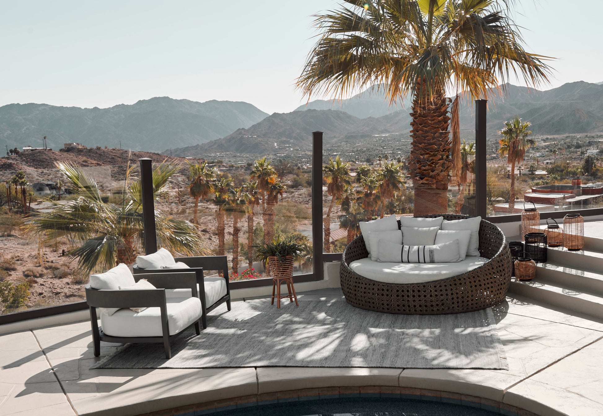 Outdoor rooftop lounge set: round dark wicker sectional with gray cushions, two white chairs, palm trees & desert mountains.