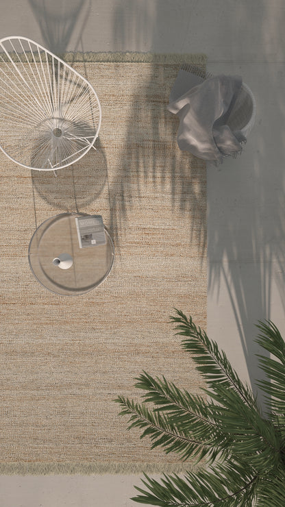 Overhead view of a beige woven rug with a white circular metal chair, a glass coffee table, gray pouf, and palm fronds.