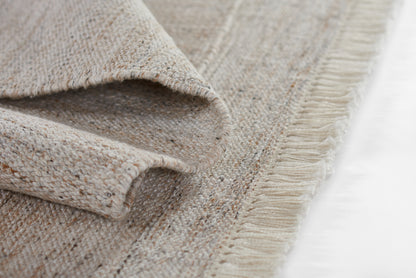 Natural beige woven rug with fringed edge and folded corner; soft textured fibers.