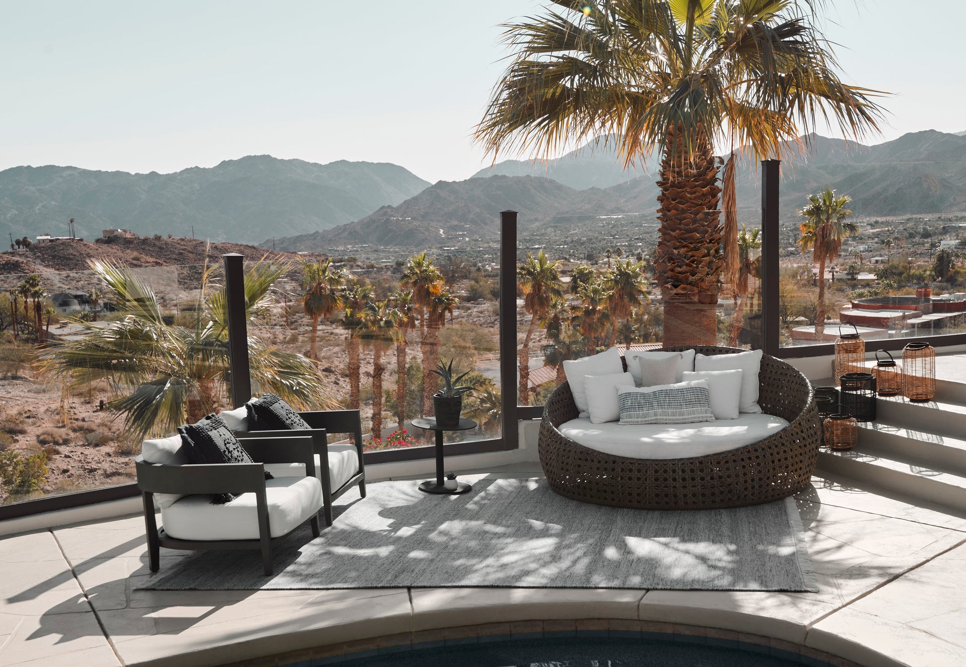 Outdoor rug 2' x 3' on a terrace with a round wicker daybed with white cushions, a black lounge chair, and palm trees with desert mountains in the background.
