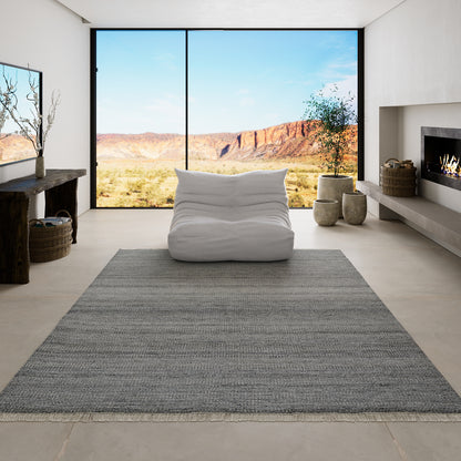 Gray woven area rug in a modern living room with a white lounge chair, desert canyon view, fireplace, and neutral decor.