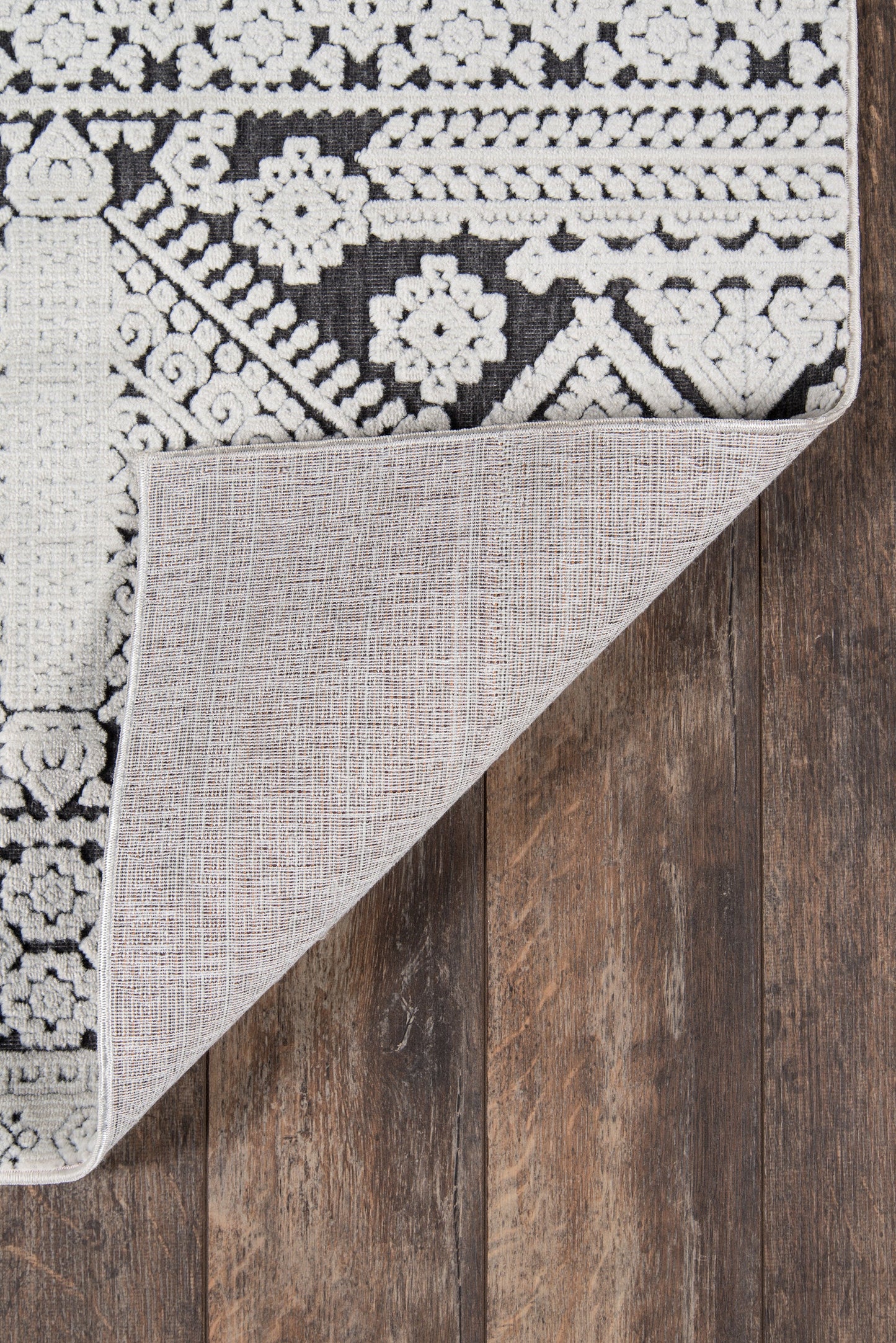 Black & white patterned area rug with floral-geometric motifs; corner folded to reveal light gray backing on dark wooden floor.