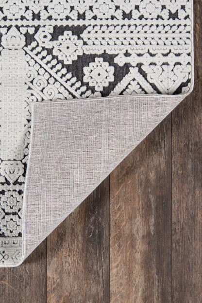 Black & white patterned area rug with floral-geometric motifs; corner folded to reveal light gray backing on dark wooden floor.