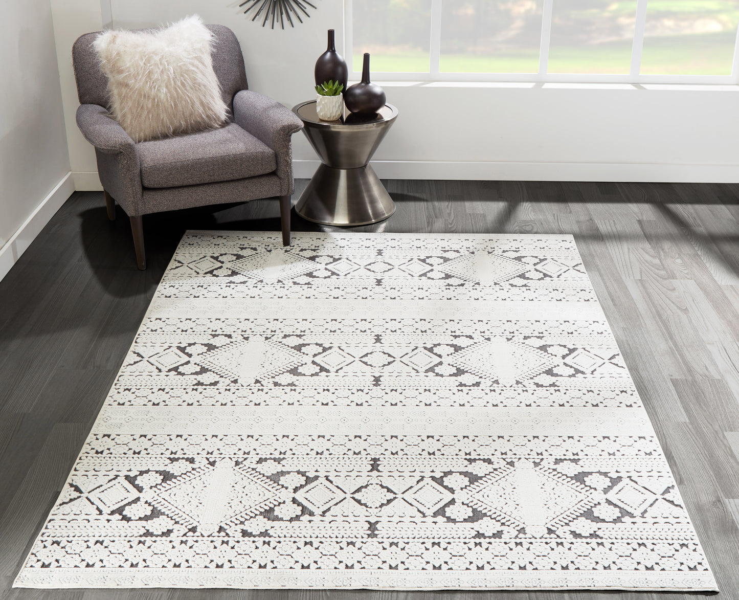 Light gray geometric rug with diamond pattern on dark wood floor, gray chair with cream fluffy pillow and metal side table.