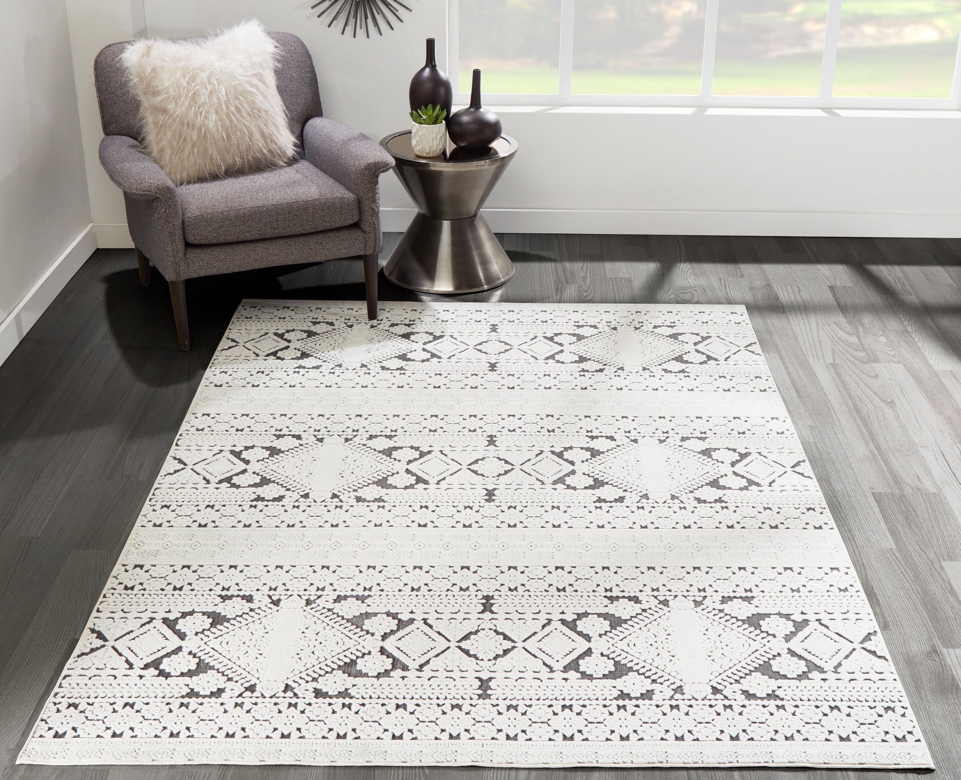 Light gray geometric rug with diamond pattern on dark wood floor, gray chair with cream fluffy pillow and metal side table.