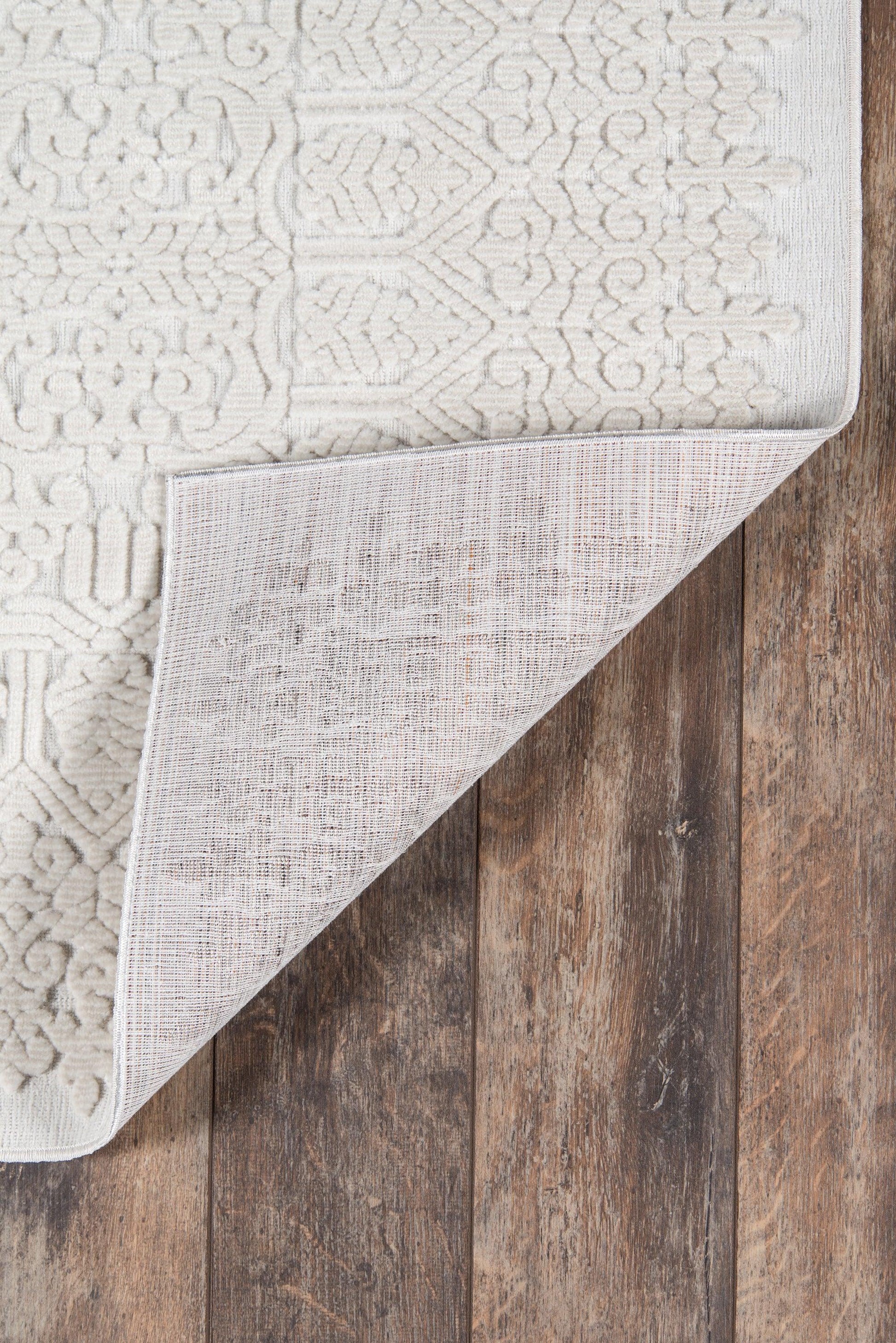 Ivory textured woven runner rug with subtle damask patterns; folded corner reveals soft backing on a dark wooden floor, 2' x 8'.
