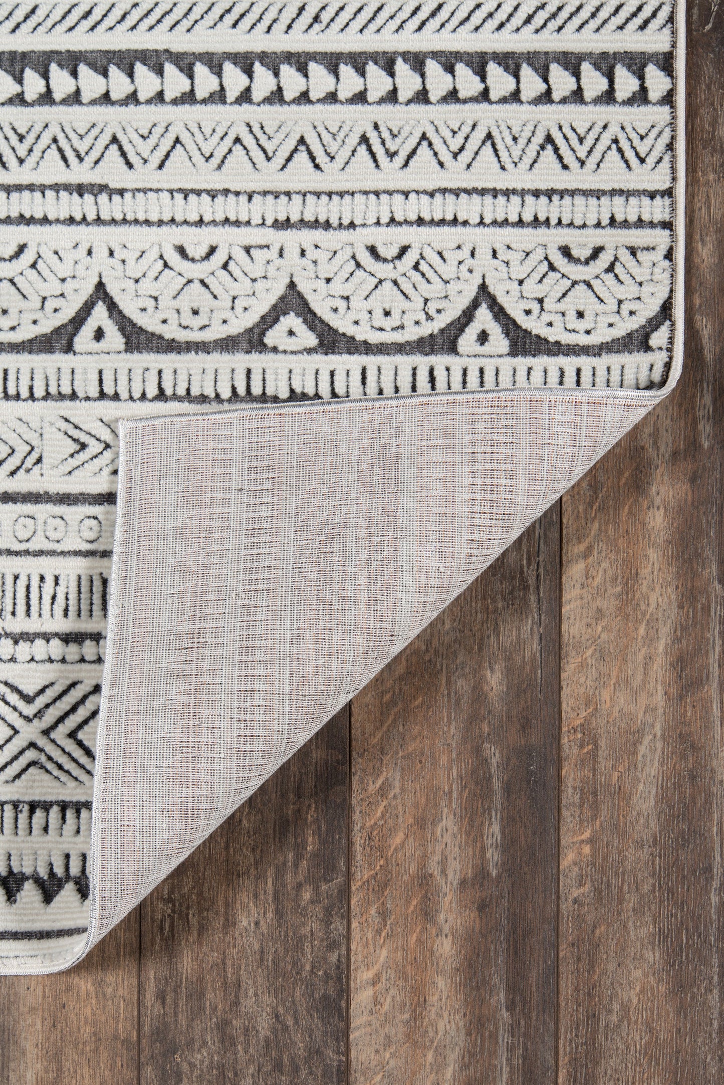 Woven black-white geometric rug with tribal motifs, folded to show beige underside, laid on wood floor.