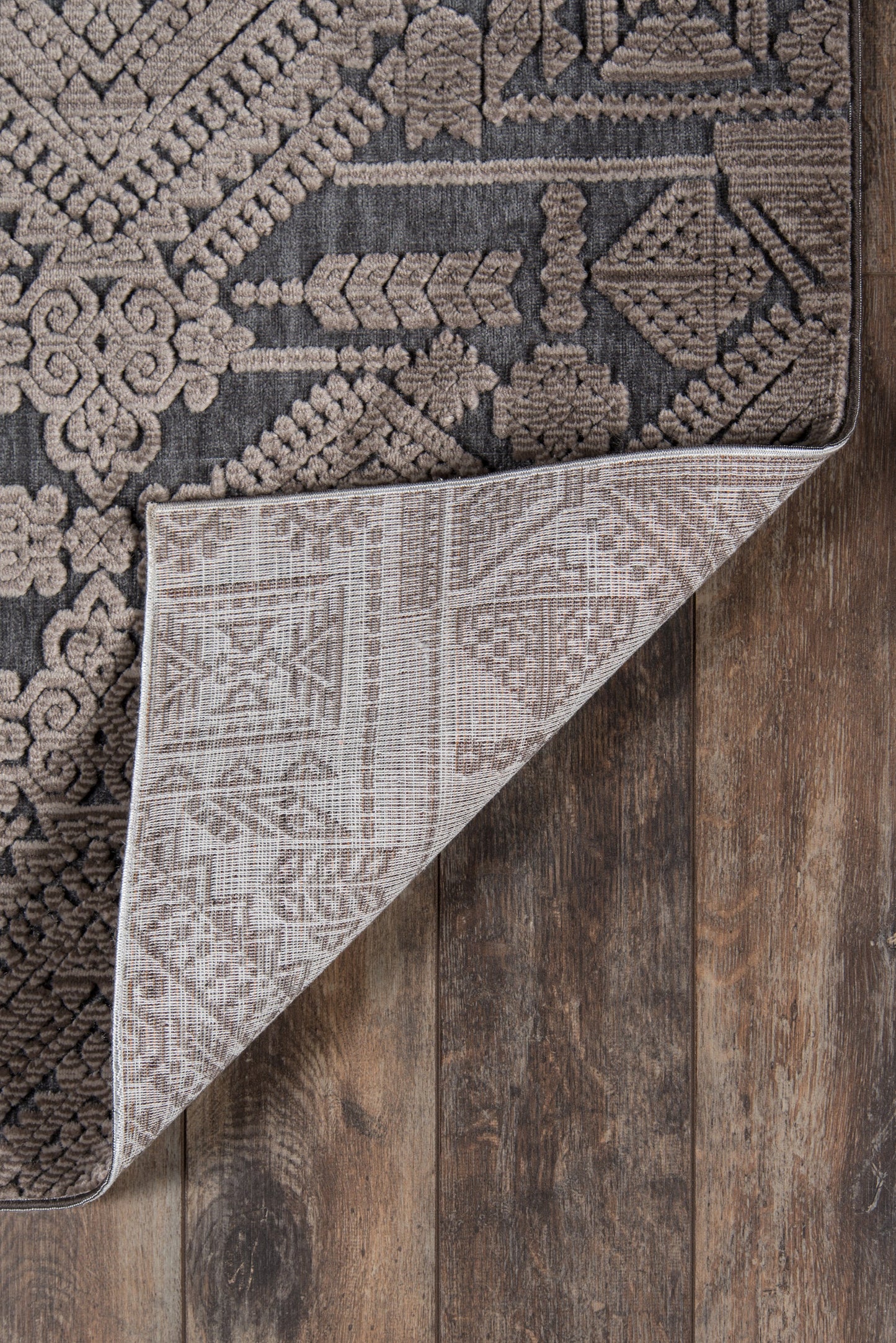Brown and taupe oriental-style woven rug with geometric motifs, folded at the corner to reveal a light beige underside on a wooden floor.