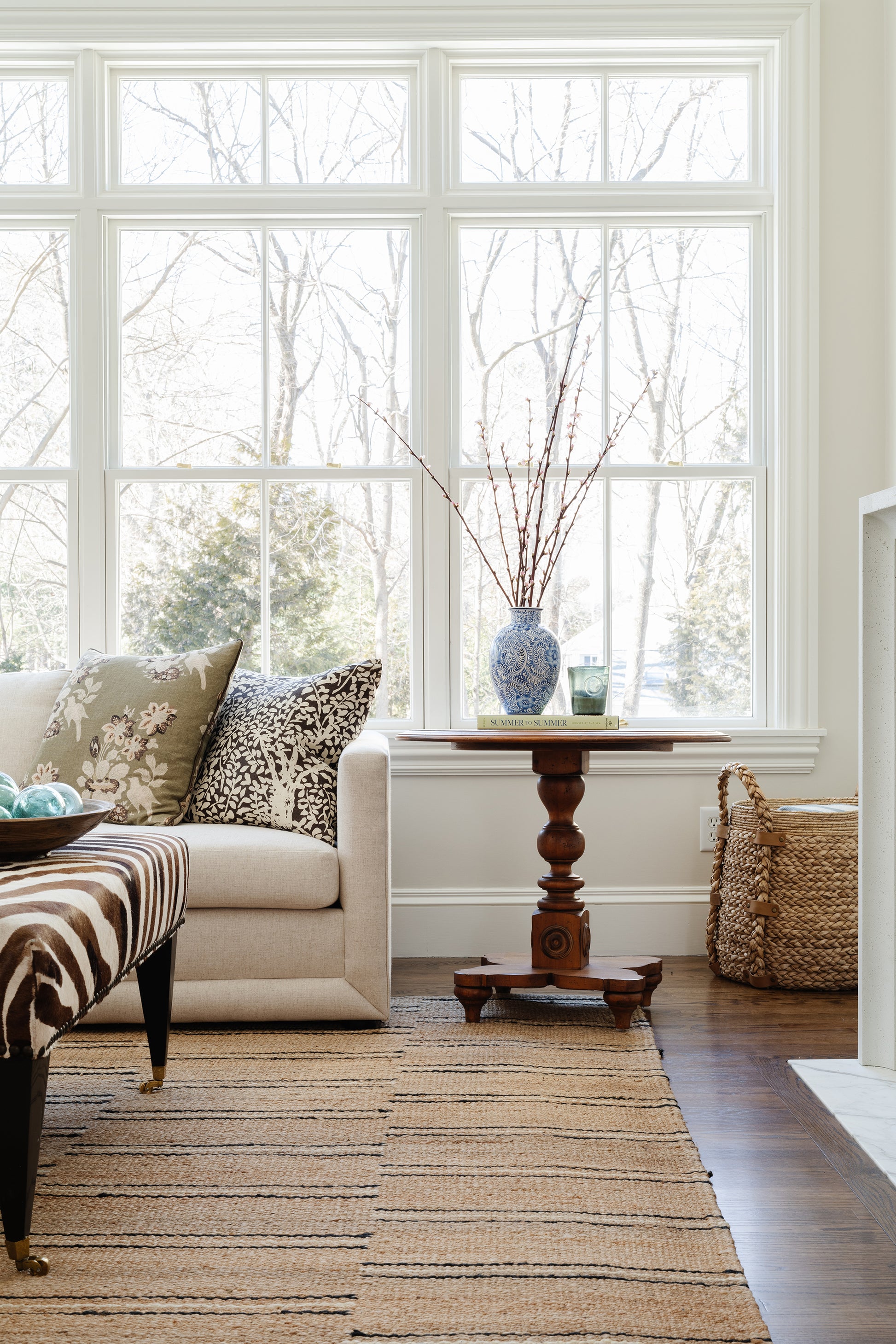 Cozy living room with beige sofa, mixed cushions, striped rug, natural wood side table, blue vase with branches, wicker basket, grid window.
