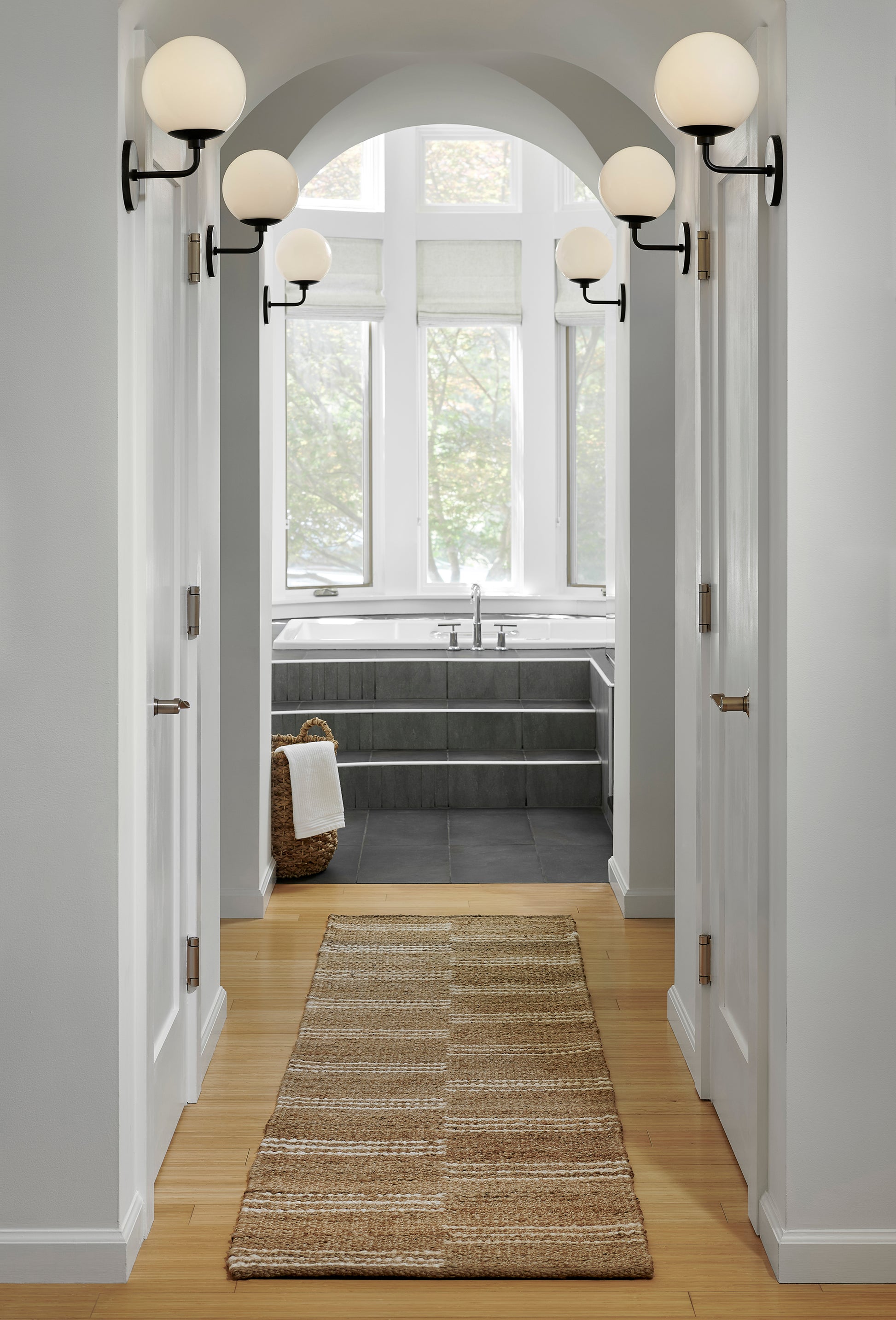 Bright white hallway with warm wood floor, arched doorway to sunlit bathroom, large window, globe wall sconces, beige runner rug.