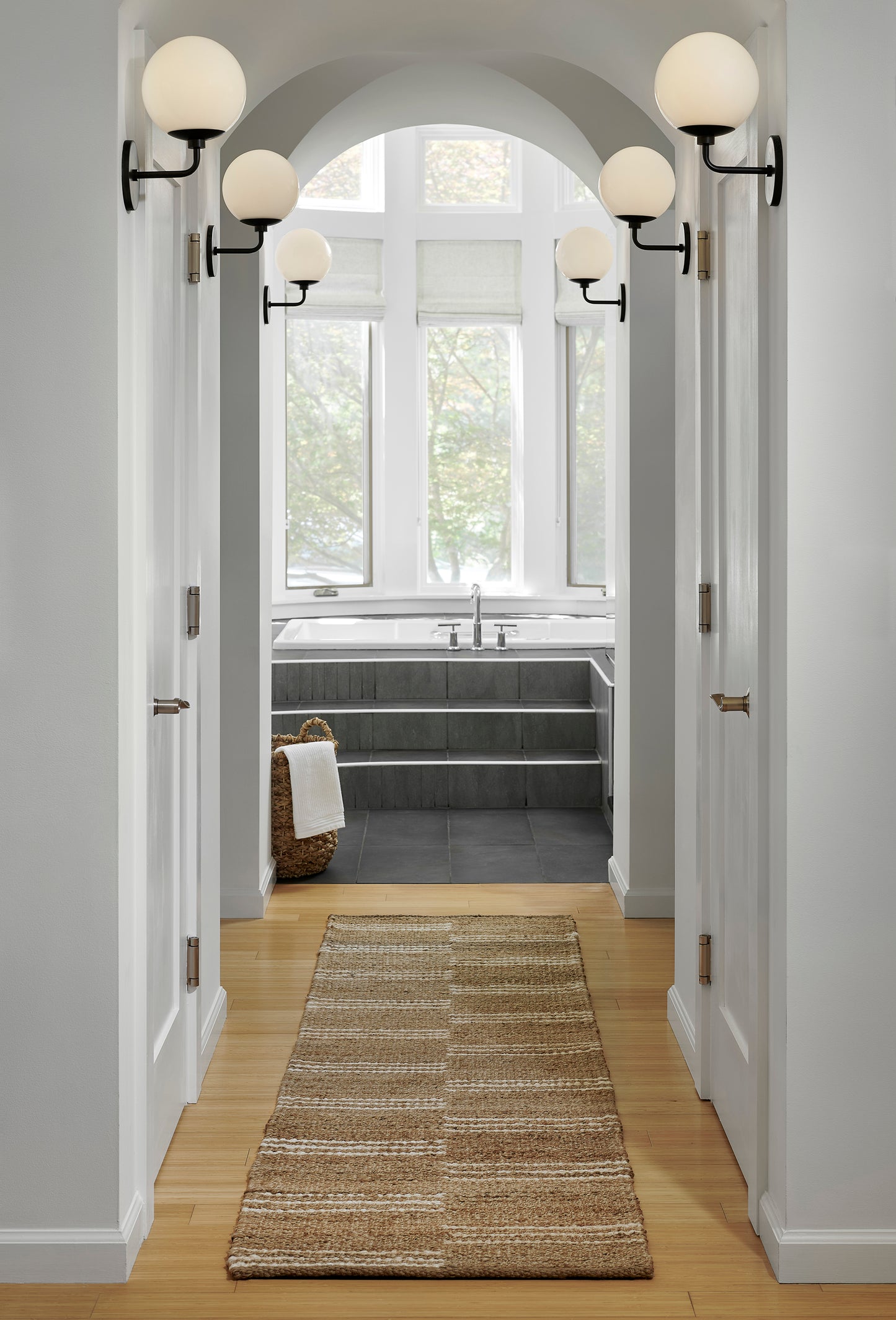 Bright white hallway with arched opening to a grey-tiled bath; black globe sconces and a light wood floor with a beige runner.