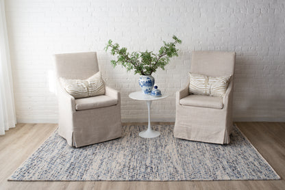 Two beige slipcovered armchairs flank a white round table with a blue vase of greenery on a blue patterned rug.
