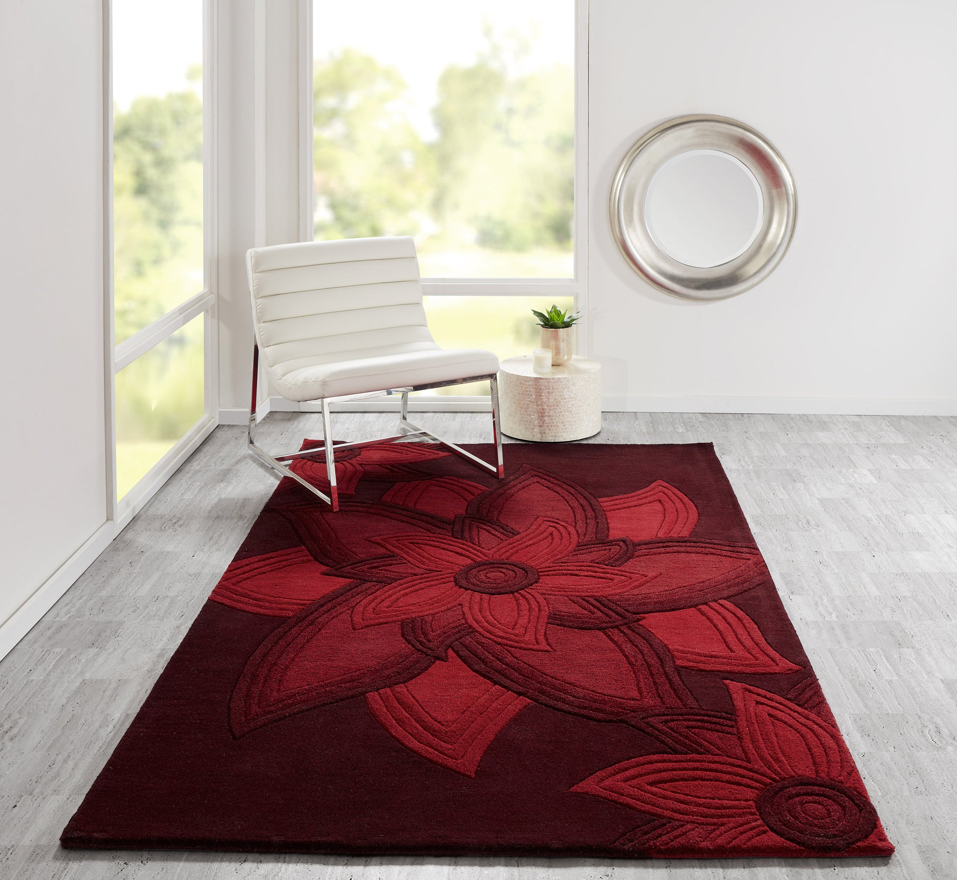 Deep red floral area rug with oversized petal motif in a sunlit room beside a white lounge chair & round wall mirror.