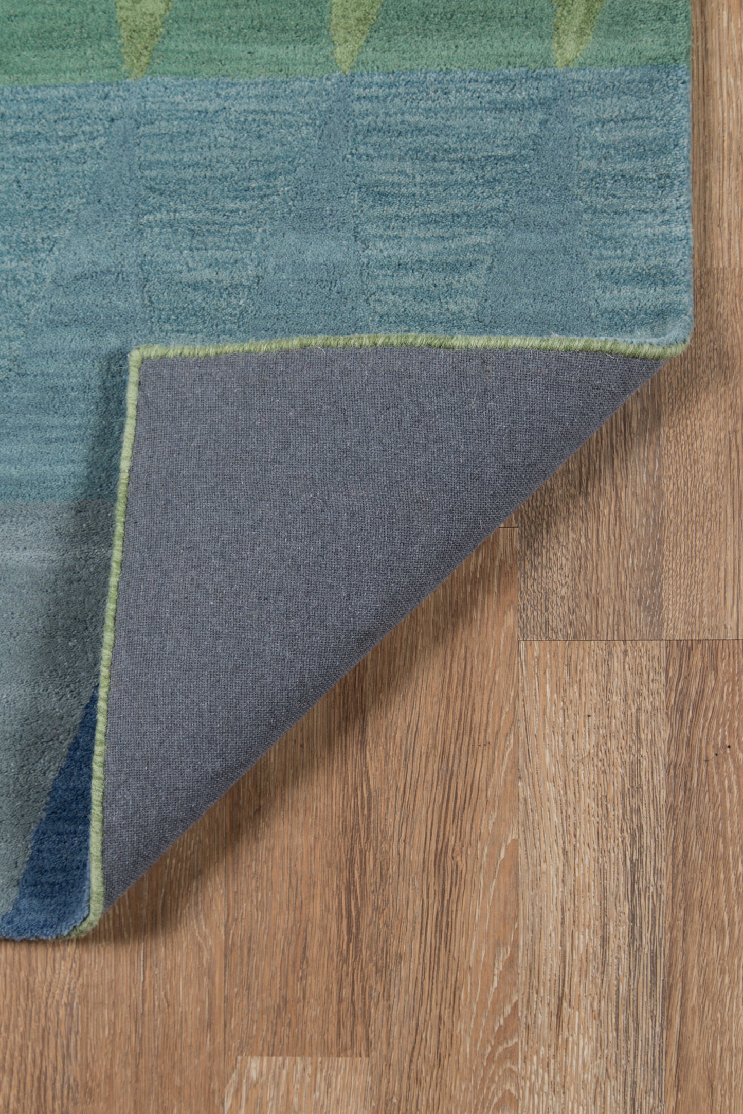 Teal and blue textured rug with lime green edge trim, folded corner revealing gray underside on wood floor.