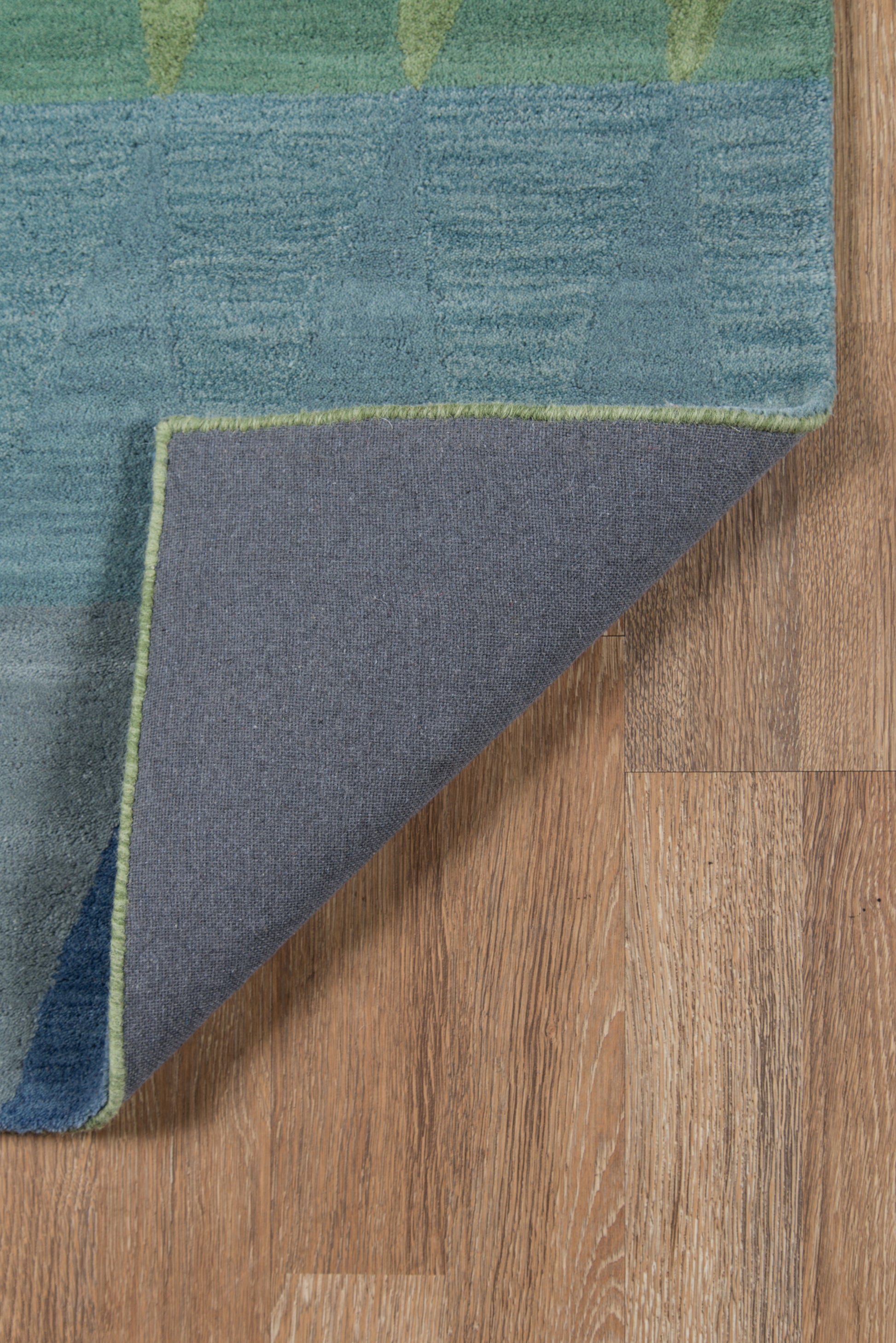 Blue-green geometric rug with lime trim; folded corner reveals gray underside on wooden floor.
