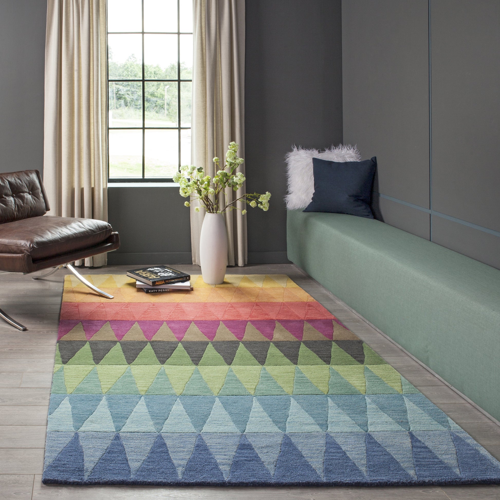 Rainbow triangle rug on wood floor with green bench, blue pillow, brown leather chair, vase of greenery by window.