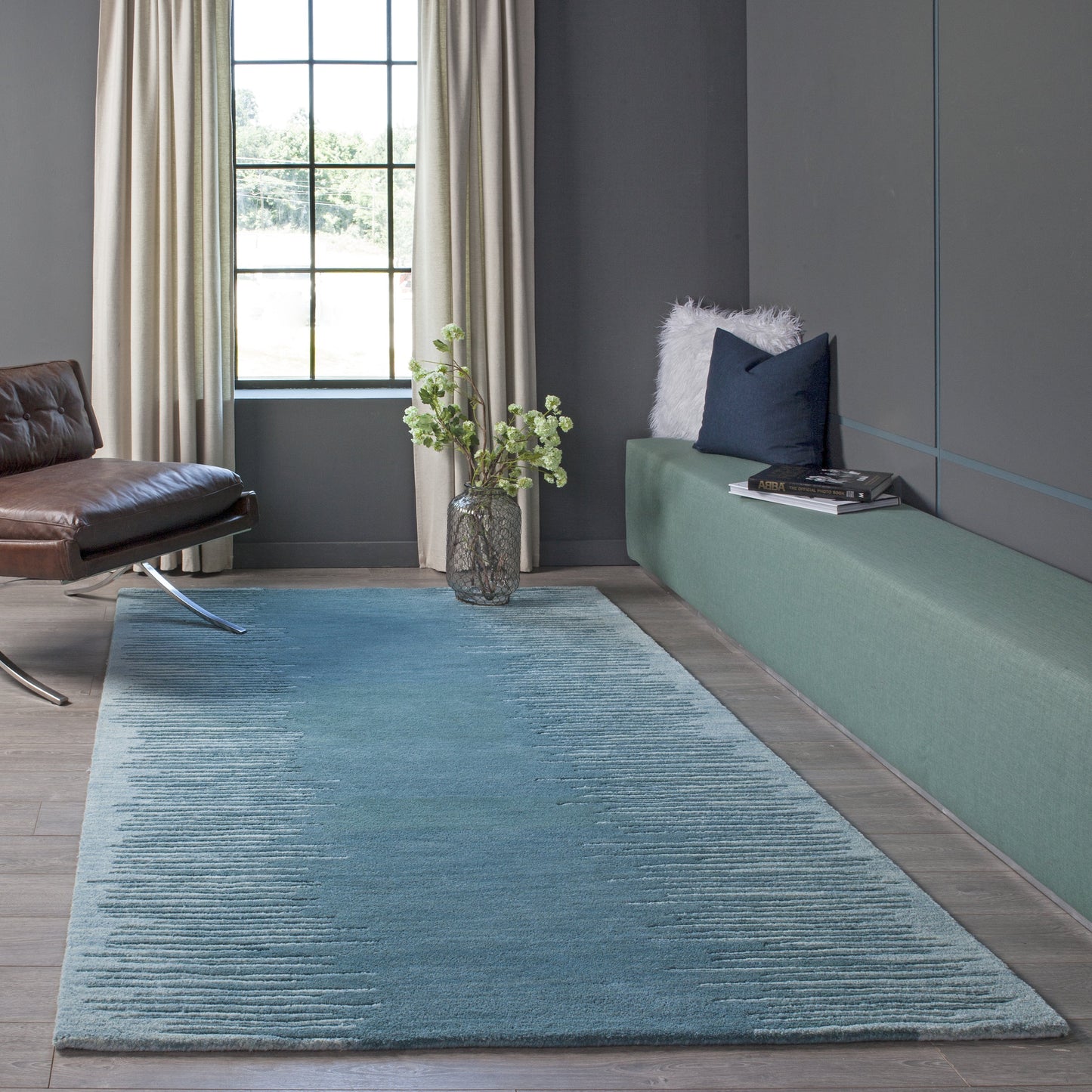 Blue textured rug in a modern living room with charcoal walls, brown leather chair, green built-in bench & window.