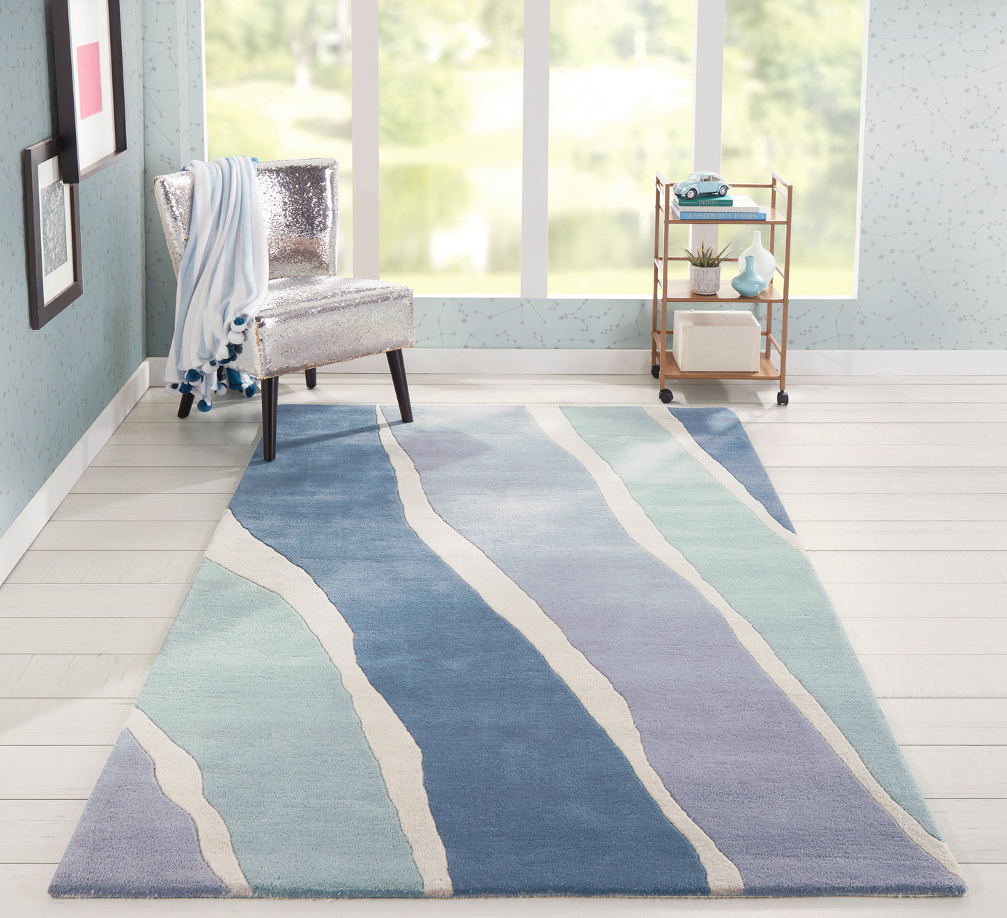 Coastal living room featuring a large blue wavy-striped area rug, a silver upholstered accent chair, and a gold bar cart.