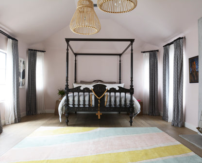 Dark wood four-poster bed with white linens, gold rosary draped on the footboard; pastel striped rug, rattan pendant lights, airy curtains.