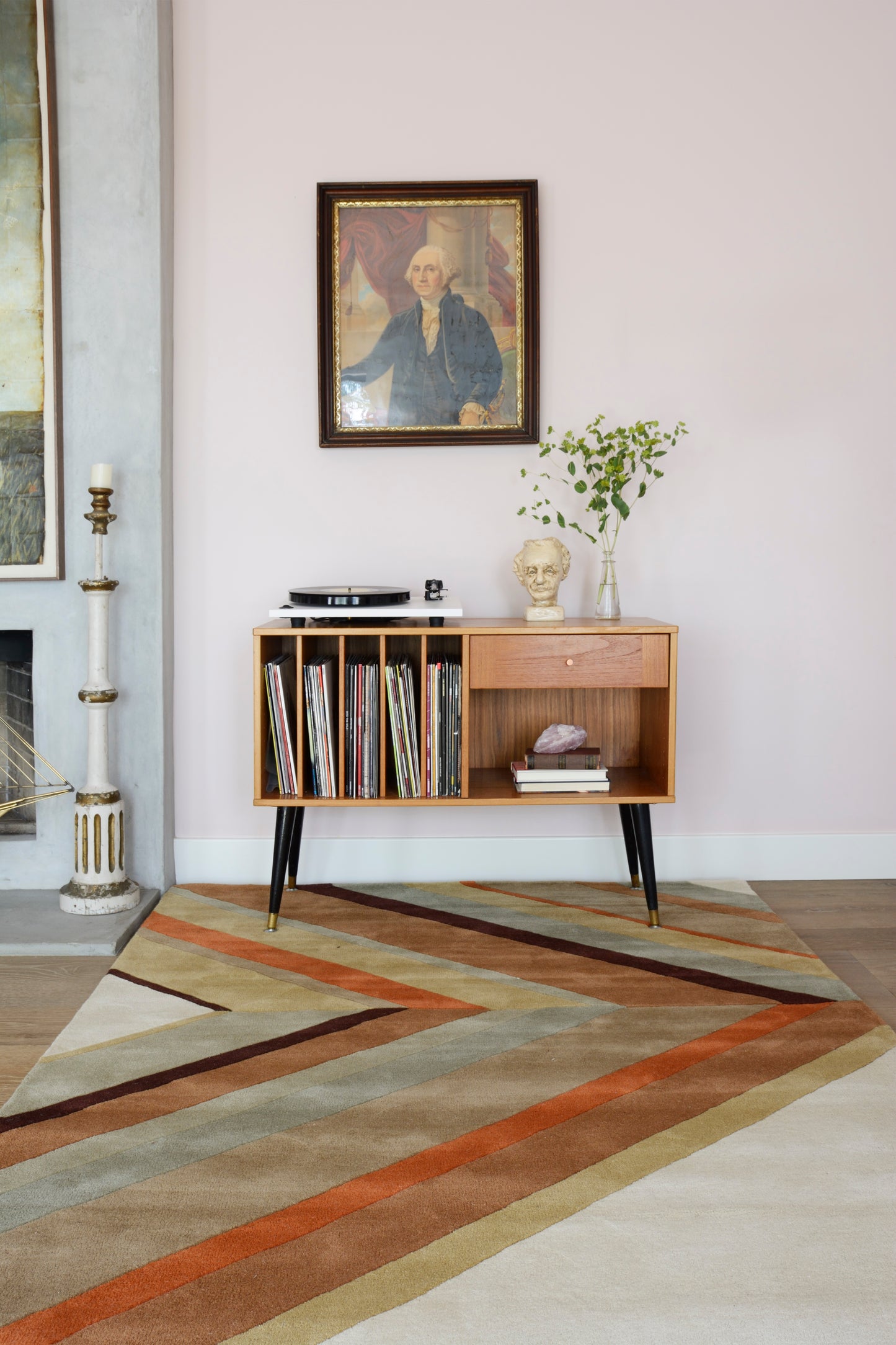 Rug in a living room with pale pink wall; mid-century teak sideboard with black legs, vinyl records and books, a bust, vase of greens, and a framed portrait above.