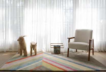 Bright living room with sheer curtains, two fluffy dogs, a light gray armchair with wooden arms, and a colorful striped rug.