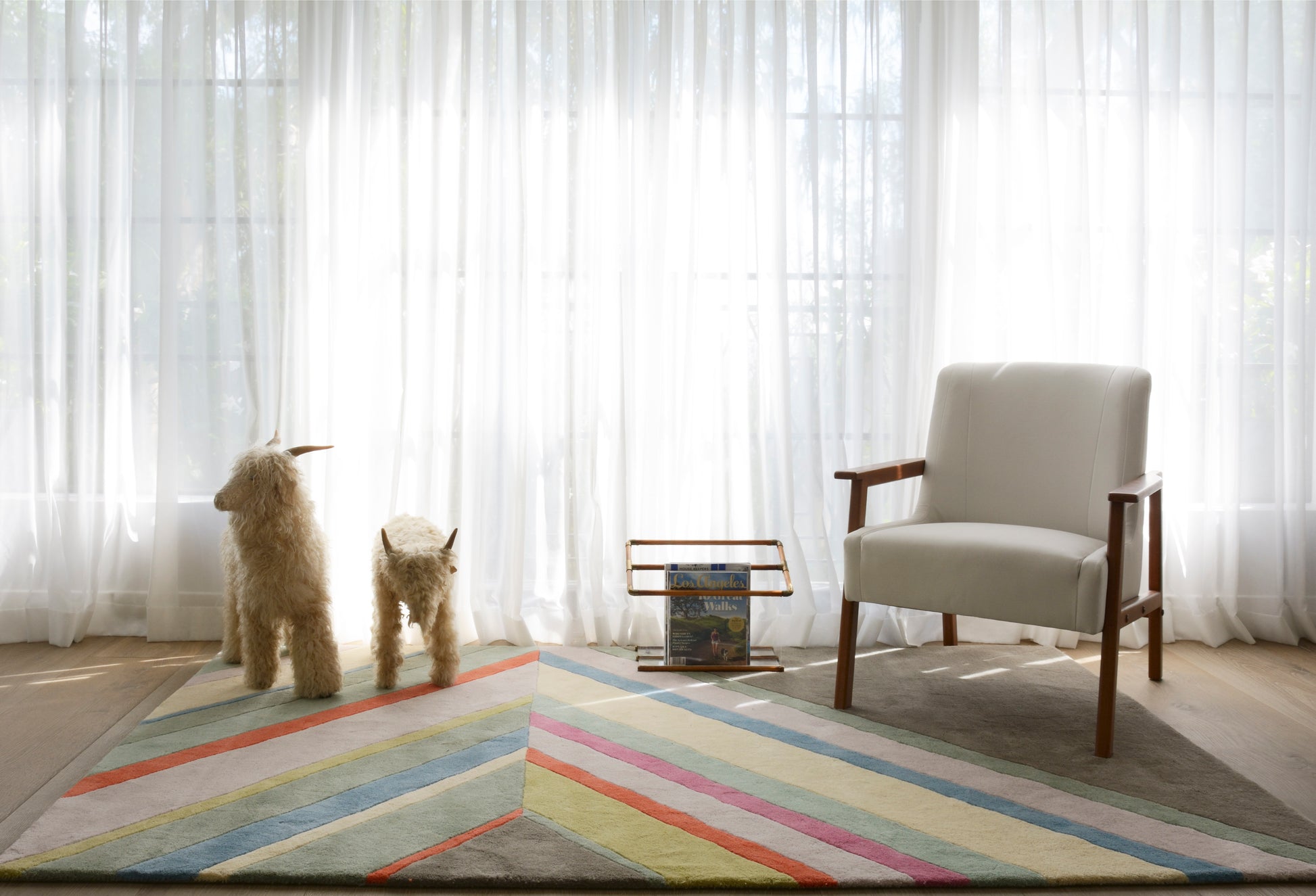 Pastel striped area rug on a sunlit living room floor with a light gray armchair, magazine rack, and two fluffy dogs.