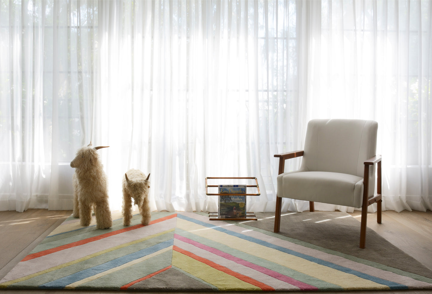 Bright living room: sheer curtains, two beige poodles, a beige armchair with wooden arms, and a multicolor striped rug.