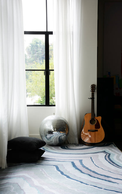 Cozy room corner with sheer white curtains, blue striped rug, silver disco ball, black cushions, and an acoustic guitar.