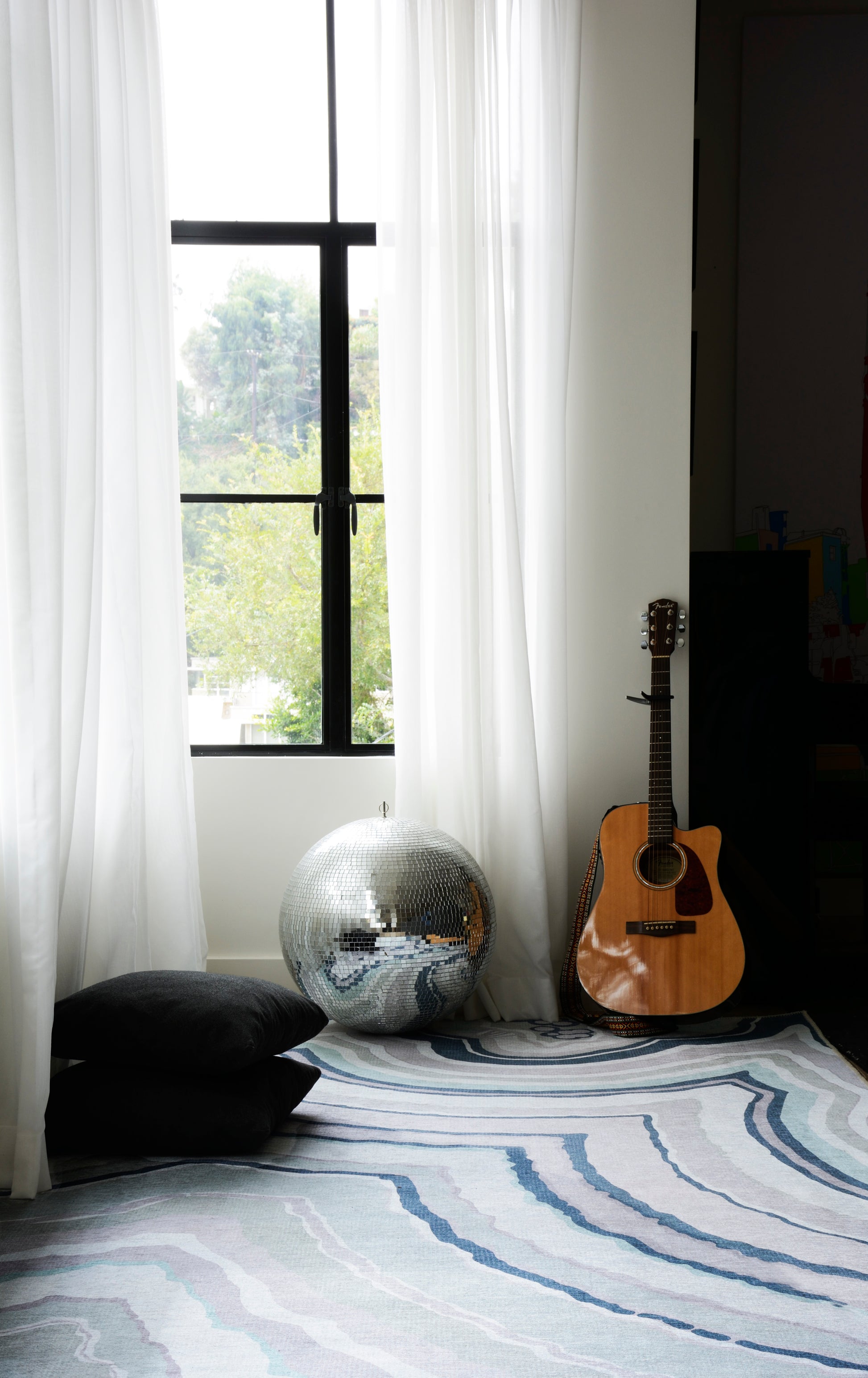 Living room corner with acoustic guitar, mirrored disco ball, blue wave rug, black floor cushions, white sheer curtains.