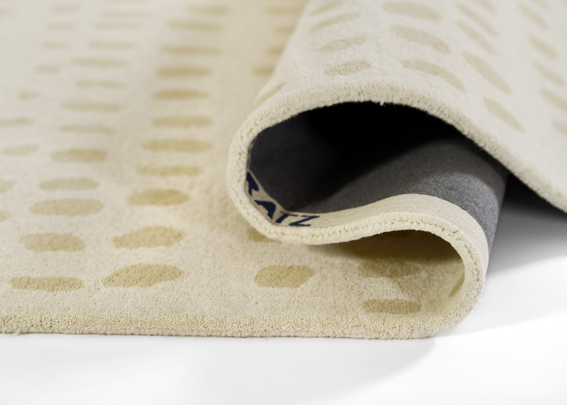 Beige patterned rug with tan rectangular motifs, rolled edge showing gray backing.