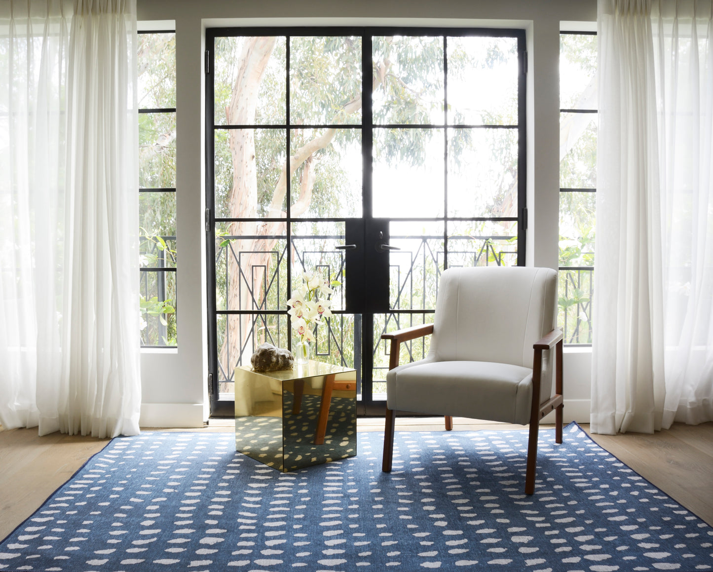 Bright living room with glass doors, sheer curtains, blue rug with white dots, cream armchair and gold cube table.