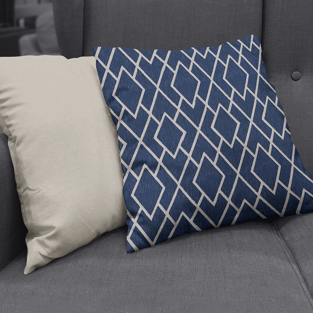 Navy diamond lattice throw pillow with white geometric lines beside a beige solid pillow on a gray sofa.