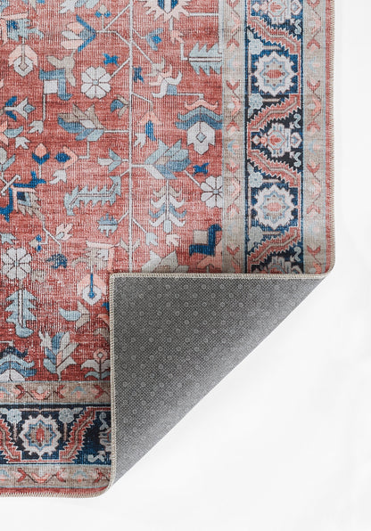 Persian-inspired pink rug with blue floral motifs and a navy geometric border; folded corner reveals grey dotted backing