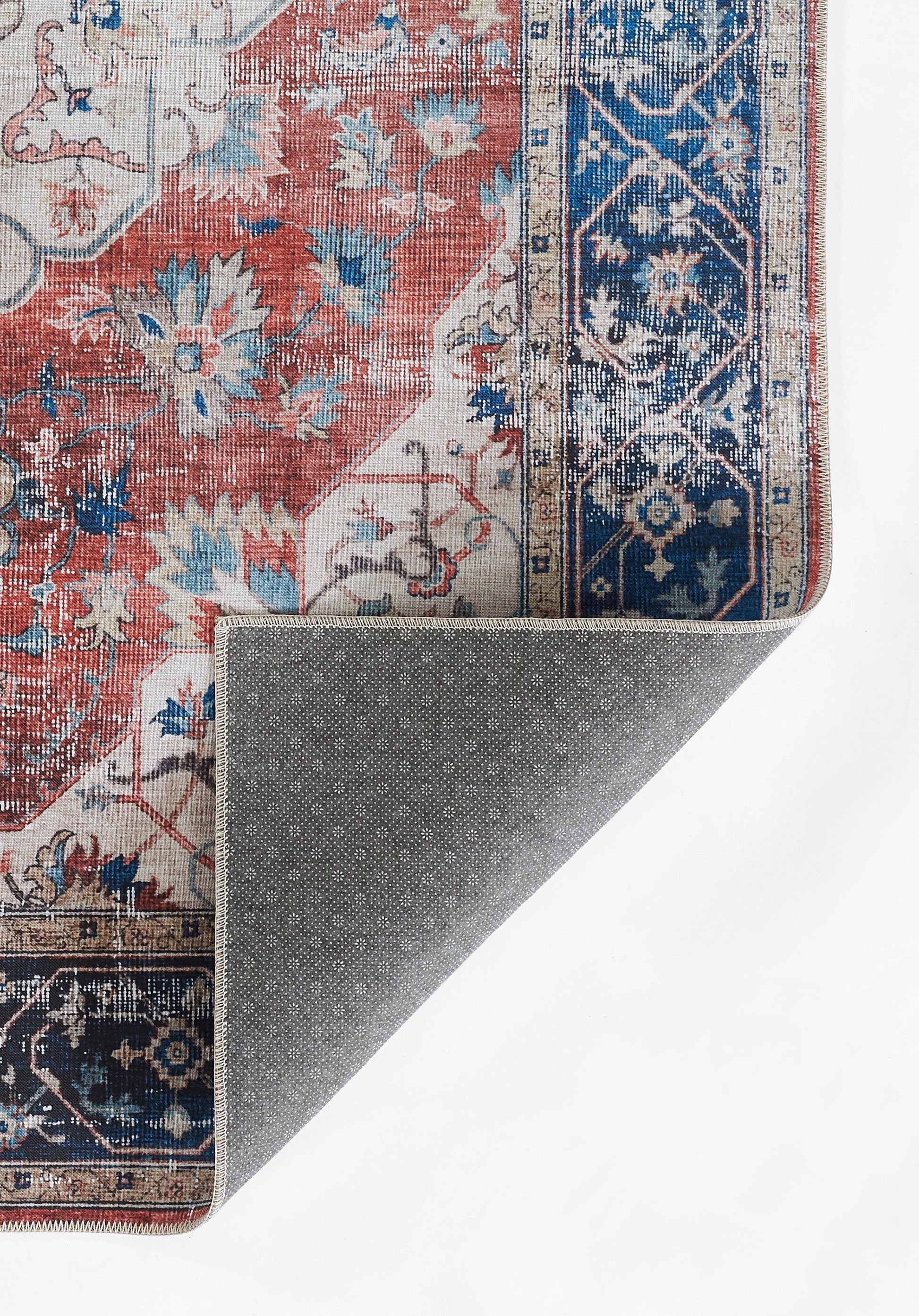 Red and blue oriental rug with floral motifs; folded corner reveals grey dotted backing.