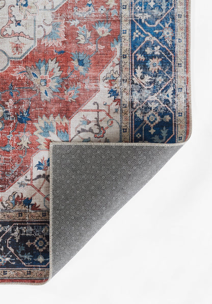 Red and blue oriental rug with floral motifs; folded corner reveals grey dotted backing.