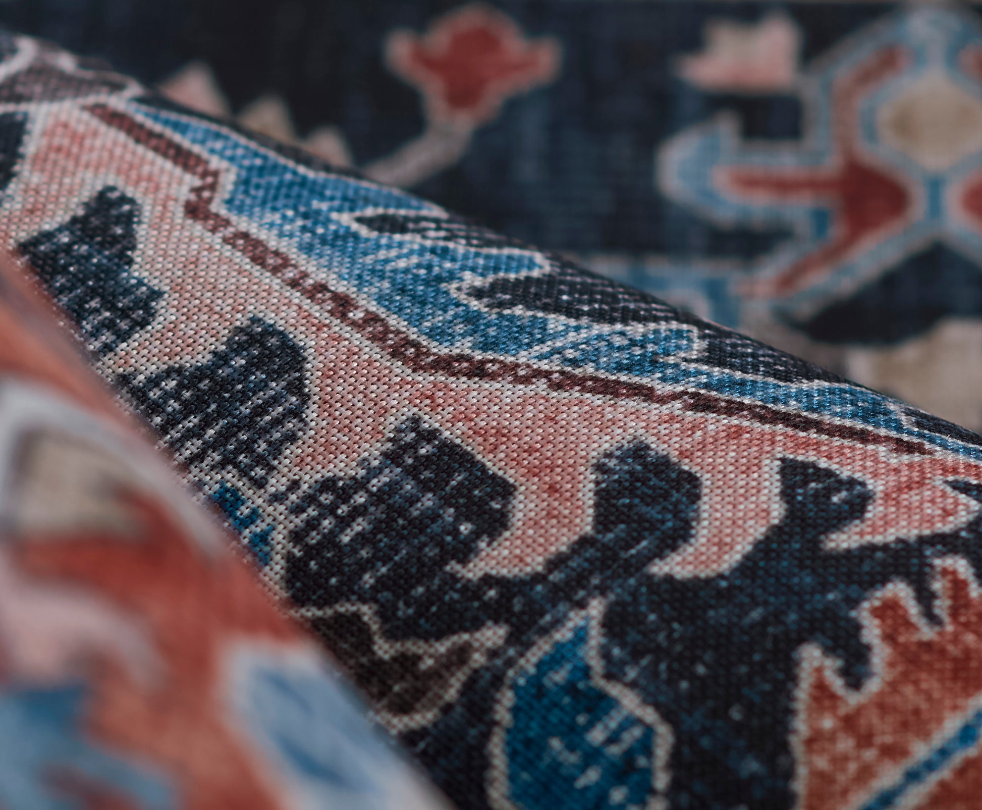 Close-up of a patterned woven rug with geometric motifs in blue, pink, teal, and cream, showing texture and folds.