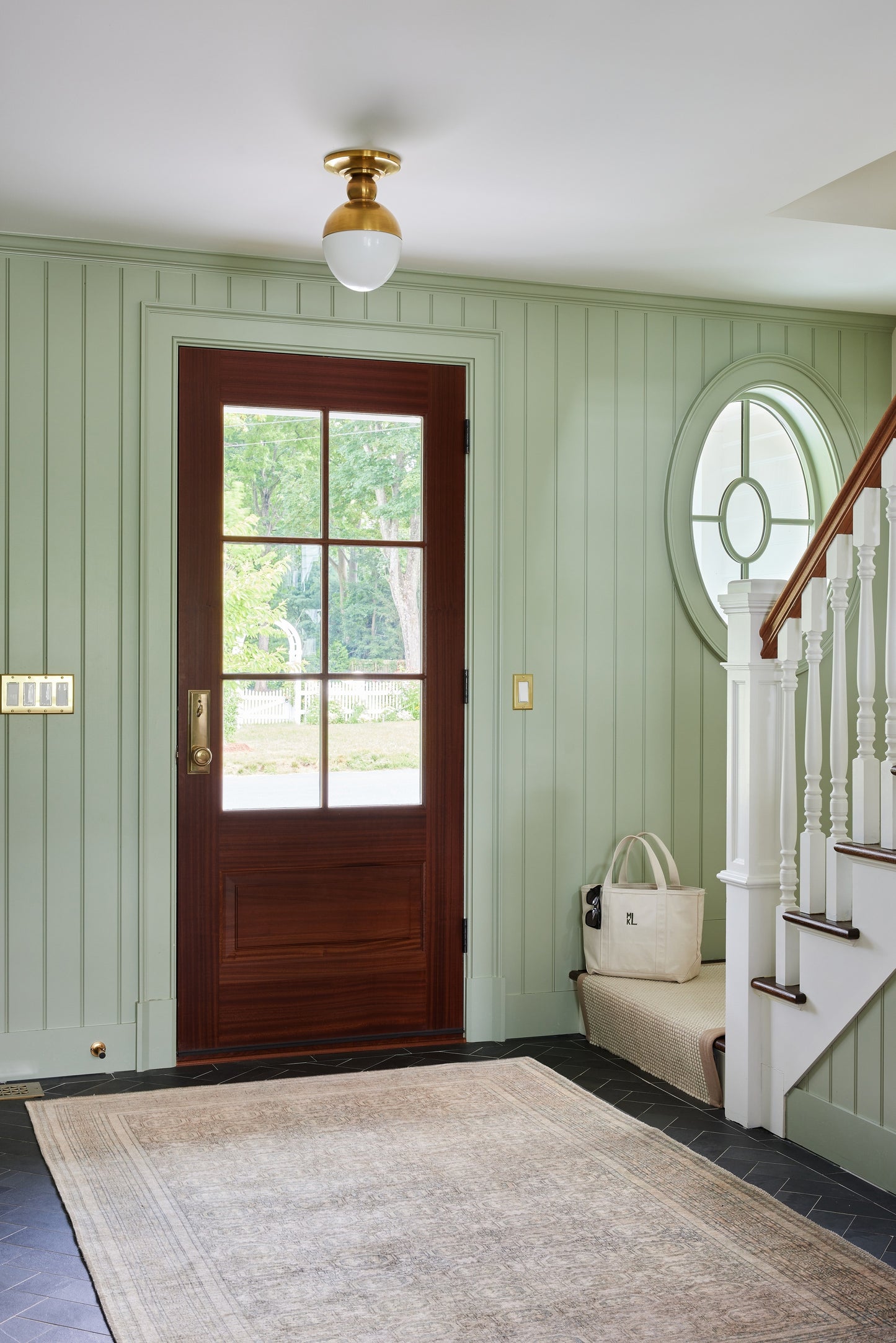 Dark wood front door with glass panes, mint green wainscoted entry, round window, staircase, beige rug and tote bag on bench.