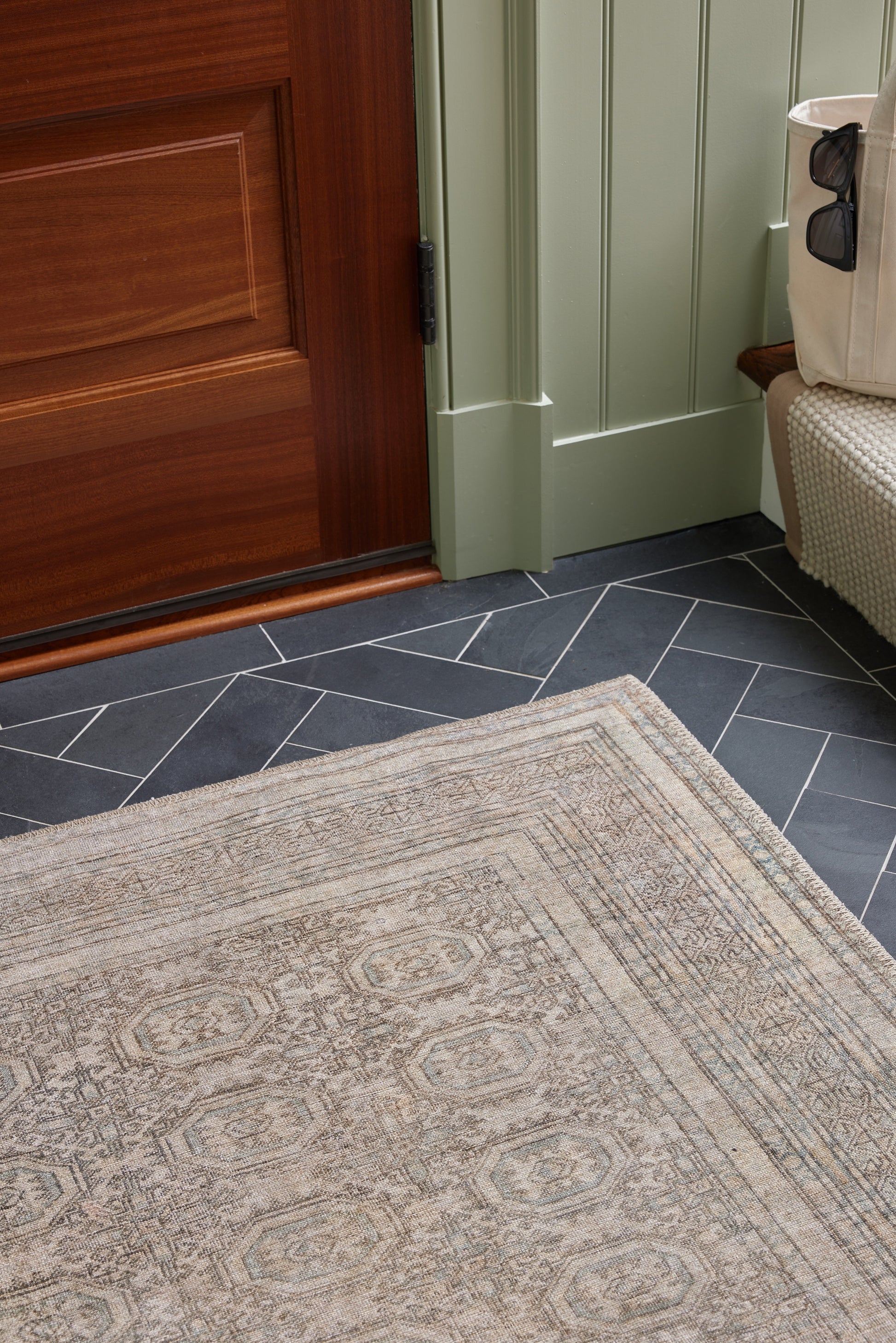 Beige-gray woven rectangular rug with geometric medallion border on dark slate tile floor beside a green paneled door.