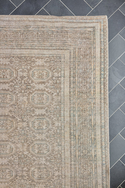 Beige oriental rug with geometric borders and repeating medallion motifs on a dark tiled floor.