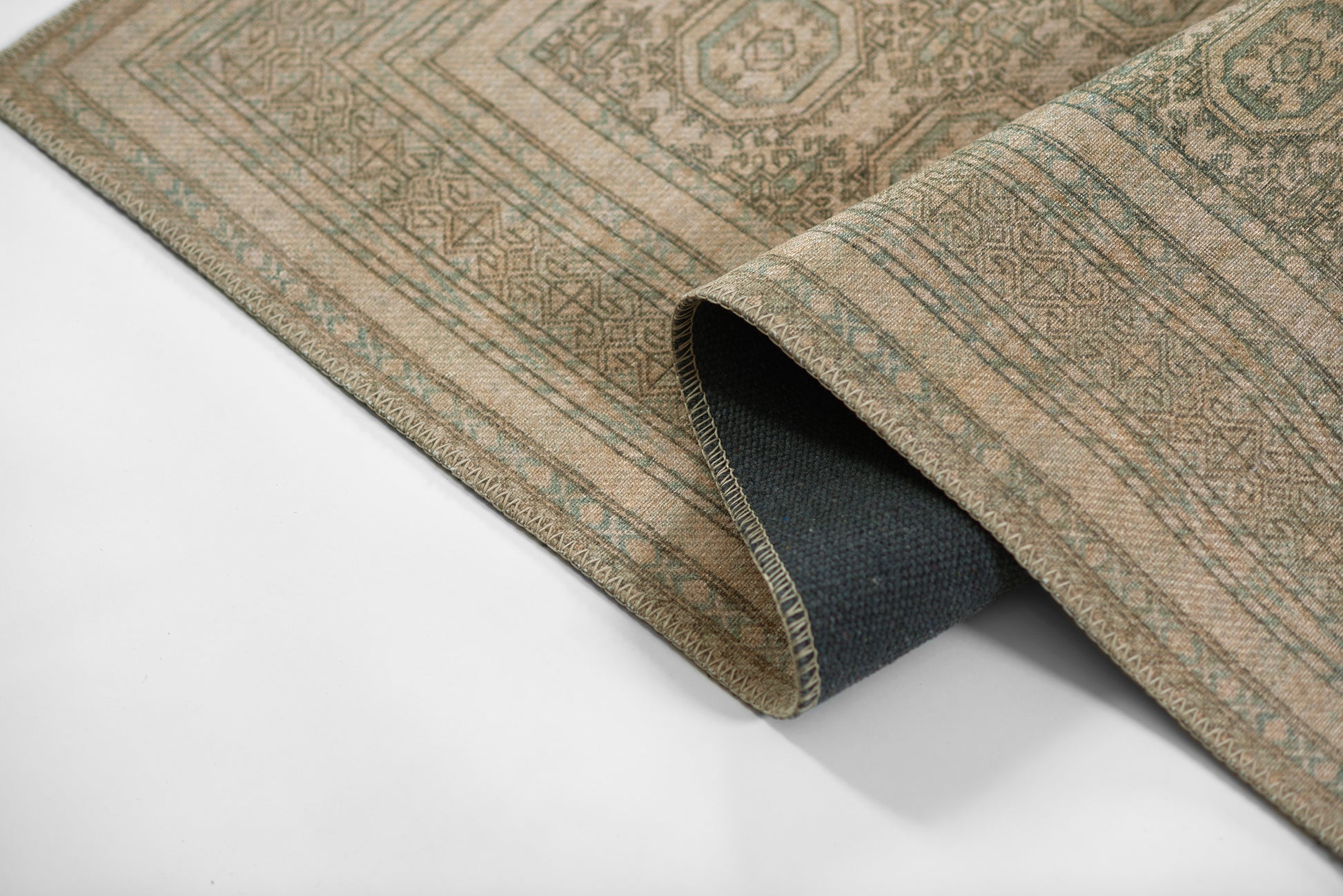 Beige woven area rug with geometric oriental motifs in sage/gray, edge rolled to show dark blue underside.