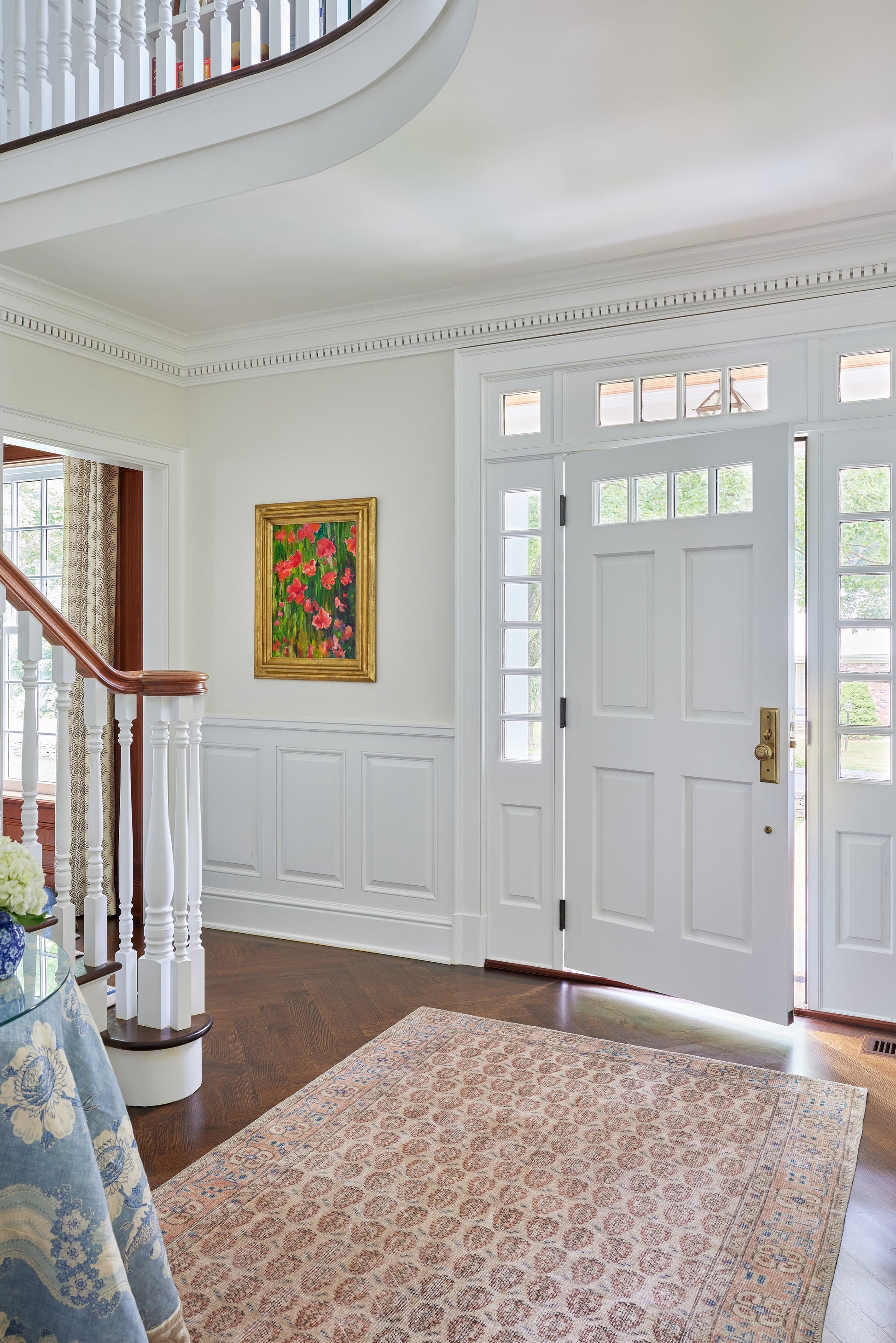 Bright white entryway with paneled front door, sidelights, transom windows, staircase, wainscoting, and a patterned area rug.