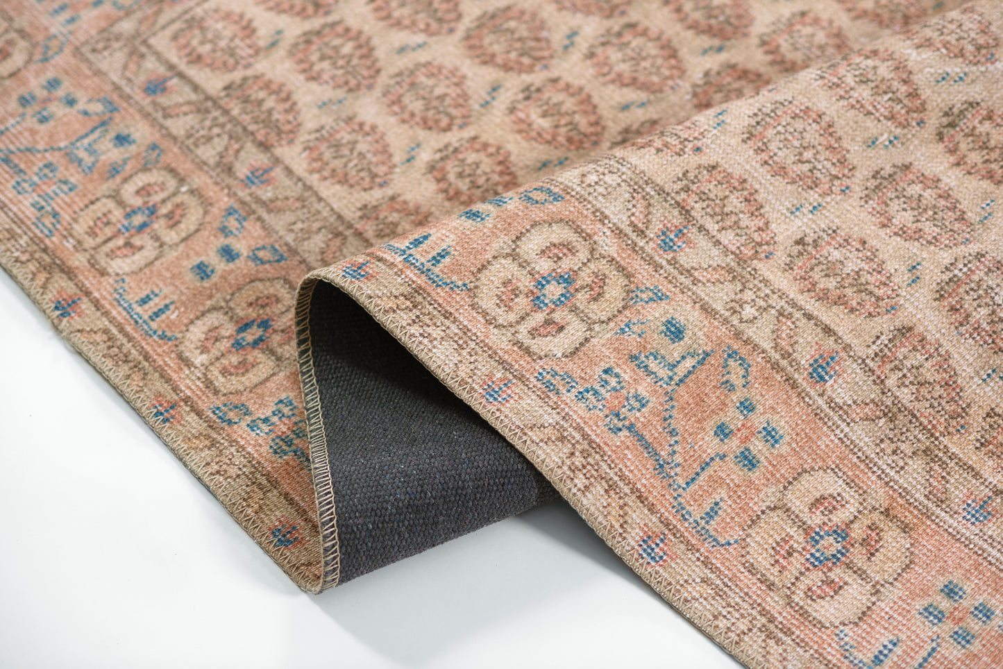 Beige-pink patterned woven rug with blue floral motifs, folded to reveal a dark backing.